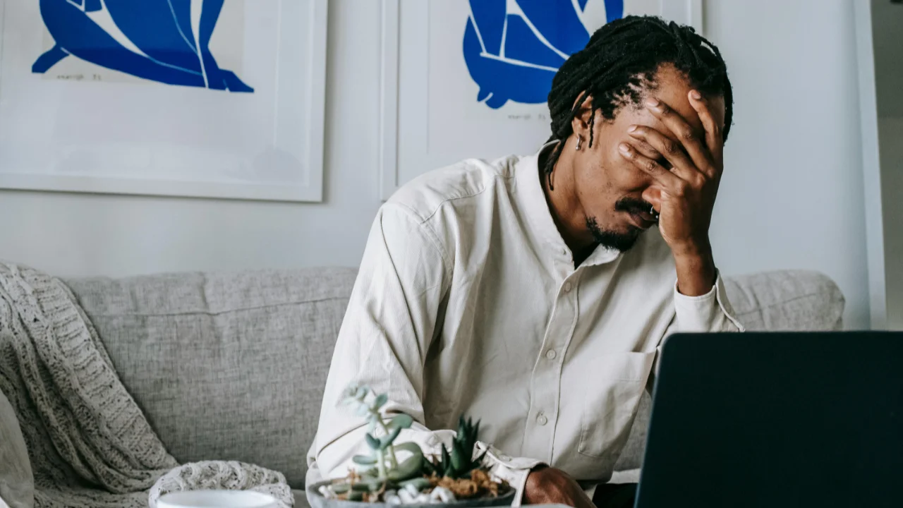 A man with dreadlocks sitting on a sofa, wearing a beige shirt, resting his head in his hand while looking at a laptop, appearing stressed or frustrated.